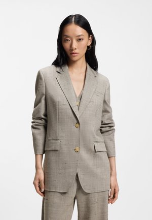 Kobieta z długimi czarnymi włosami, ubrana w beżową, teksturowaną marynarkę na dwa guziki oraz pasujące spodnie, na jednolitym białym tle.