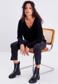 Schwarzer gerippter V-Ausschnitt-Pullover, schwarze verkürzte Jeans mit ausgefransten Säumen und schwarze Leder-Stiefeletten mit dicken Sohlen, auf einem Holzstuhl sitzend.