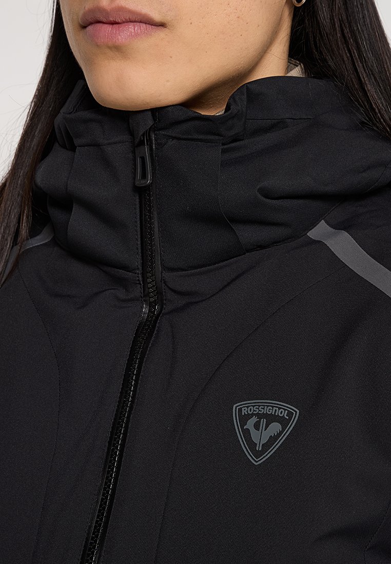 Frau trägt eine schwarze Rossignol-Jacke, bis zum Hals zugezippt, zeigt das Markenlogo und einen hohen Kragen mit reflektierendem Streifen auf der Schulter.
