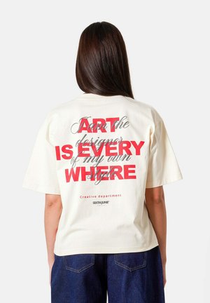Femme aux longs cheveux foncés portant un T-shirt blanc avec un texte superposé rouge et noir au dos, et un jean bleu foncé.