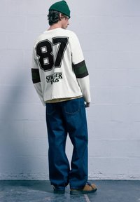 Biele tričko s dlhým rukávom a čiernymi a zelenými pruhmi, na chrbte má veľké "87". V outfite sú aj modré voľné džínsy a hnedé topánky.