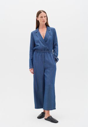 Tuta in denim con design a portafoglio, maniche lunghe e vita cinchata. Colore blu intenso, taglio a gamba larga e dettagli di cucitura sottili.