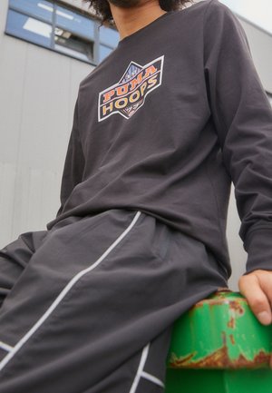 Langärmliges graues Shirt mit "PUMA HOOPS"-Logo; kombiniert mit grauen Hosen mit weißen Seitenstreifen. Modell sitzt auf einer grünen Fläche.