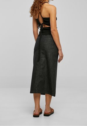 Mujer con cabello rizado que lleva un top negro de halter con lazos en la espalda, una falda midi de mezclilla oscura de cintura alta y sandalias marrones, de pie sobre un suelo gris.