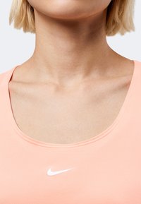 Γυναίκα που φοράει ροδακινί μπλουζάκι με λαιμόκοψη και λευκό λογότυπο Nike στο κέντρο κάτω από τη λαιμόκοψη, εμφανίζοντας το λαιμό και τις κλείδες.