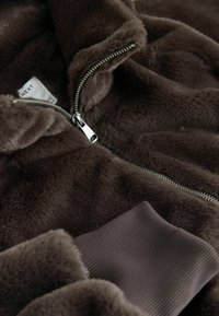 Jachetă din blană artificială maro, cu fermoar argintiu, manșete cu dungi și textură moale. Eticheta spune "NEXT."