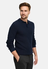 Navyblauer strukturierter Polopullover mit Kragen, langen Ärmeln und gerippten Bündchen, kombiniert mit dunkelgrauen Hosen.