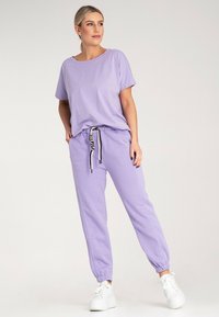 Liliowy zestaw dresowy składający się z krótkiego topu i dopasowanych spodni dresowych. Spodnie mają elastyczny pas i sznurek w paski. Białe sneakersy.