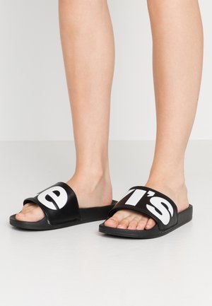 Pieds portant des sandales slides noires avec de grandes lettres blanches « e » à gauche et « t's » à droite sur un fond clair uni.