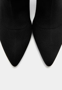 Bottines noires à bout pointu en tissu texturé, présentant un design épuré sans ferrures ni ornements visibles.