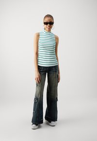 Top sin mangas de rayas verdes con cuello alto, combinado con pantalones cargo de mezclilla anchos y desgastados que cuentan con múltiples bolsillos. Zapatillas blancas.
