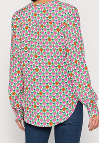 Donna che indossa una blusa a maniche lunghe con un motivo geometrico a diamante colorato in rosa, rosso, verde, blu e bianco, abbinata a jeans blu scuro.
