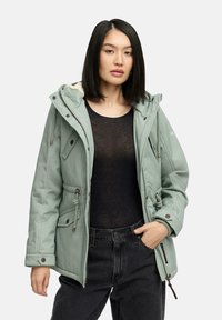Light green, insulated parka with a hood, drawstring waist, and multiple pockets. Warm lining and zipper accents. Black top and dark pants.
