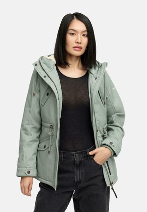 Lichtgroene, geïsoleerde parka met een capuchon, trekkoord in de taille en meerdere zakken. Warme voering en ritsaccenten. Zwarte top en donkere broek.