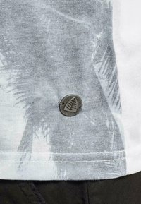 Helehall t-särk palmivihmle mustriga, millel on ümar metalltags, millel on lehekujuline disain, õmmeldud küljeserva.