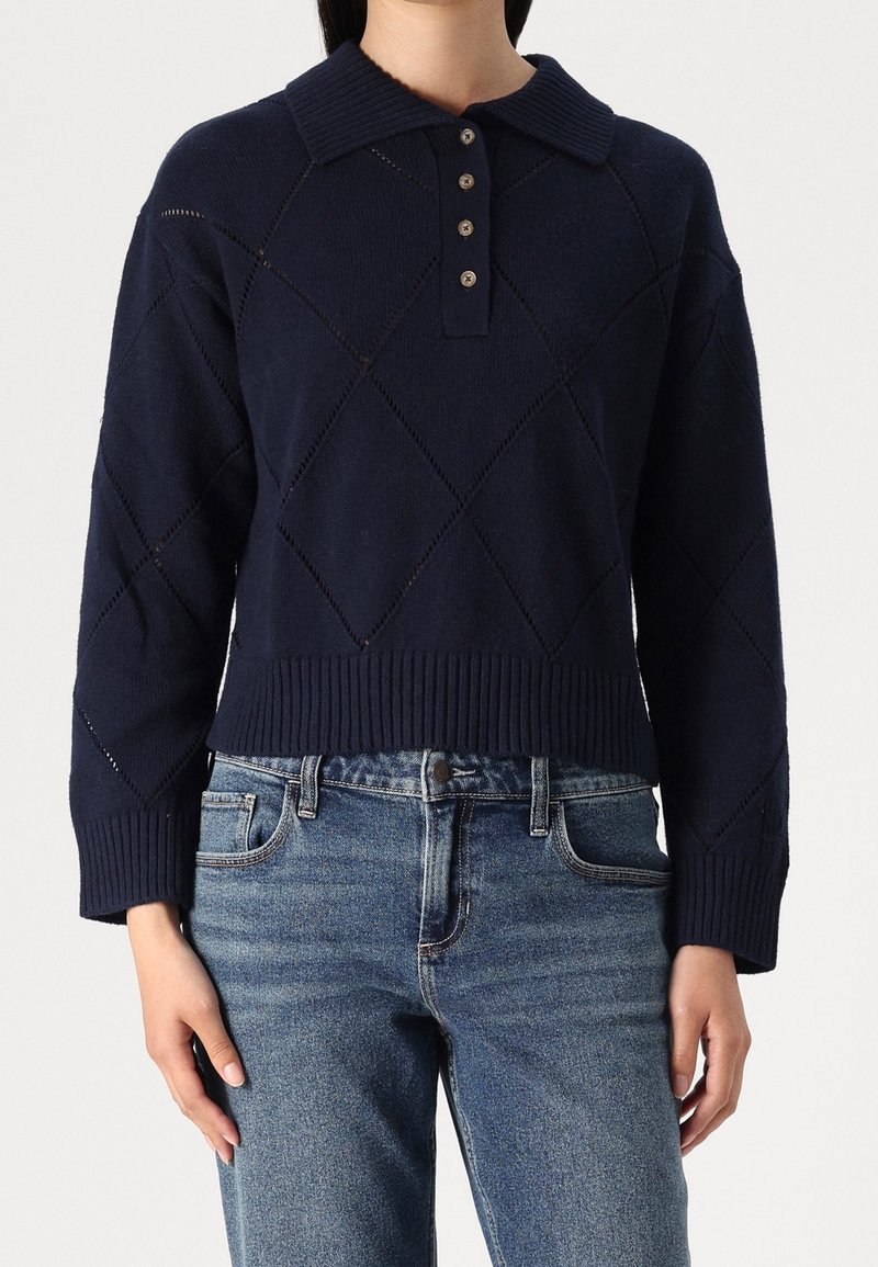 Frau trägt einen navyblauen Strickpullover mit Rautenmuster und kurzem Kragen mit Knöpfen, kombiniert mit blauen Jeans.