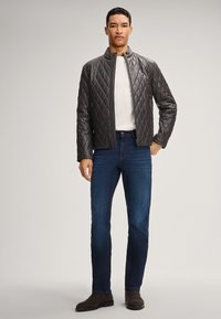 Mann in einer dunkelbraunen gesteppte Lederjacke, weißem Hemd, dunkelblauen Jeans und braunen Wildlederstiefeln, der vor einem einfarbigen Hintergrund steht.