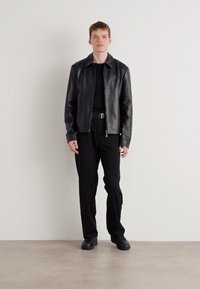 Black leather jacket with a zip front, worn over a black shirt. Paired with black trousers and shoes, all against a neutral background.