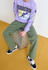 Osoba siedząca na srebrnej walizce, ubrana w fioletową koszulę z długim rękawem z grafiką, oliwkowe spodnie i czarno-białe sneakersy, na musztardowej podłodze.