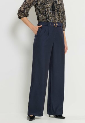 Frau trägt marineblaue, weit geschnittene, hoch taillierte Hosen mit Gürtel, kombiniert mit einer gemusterten Bluse und schwarzen, spitz zulaufenden High Heels.