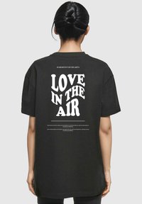 Black oversized t-shirt featuring bold white text "LOVE IN THE AIR" on the back with a subtle printed design above and below.