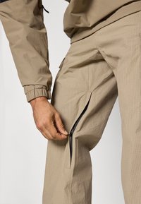 Μπεζ cargo παντελόνι με υφή, διαθέτει πλαϊνό φερμουάρ. Ένα χέρι τραβάει το φερμουάρ για ρύθμιση.