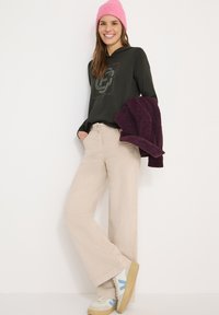 Ciemnozielona bluza z kapturem i długim rękawem z nadrukiem graficznym, beżowe spodnie o szerokich nogawkach, kremowo-niebieskie sneakersy oraz różowa dziergana czapka.