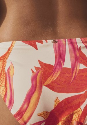 Prim-plan cu piele maro și talie purtând un costum de baie alb cu modele florale abstracte portocalii și roz și o mică închizătoare metalică.