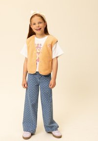 Oranje fleecevest met drukknopen over een wit T-shirt met print, gecombineerd met blauwe jeans met wijde pijpen en witte hartjespatronen, en lichte sneakers.