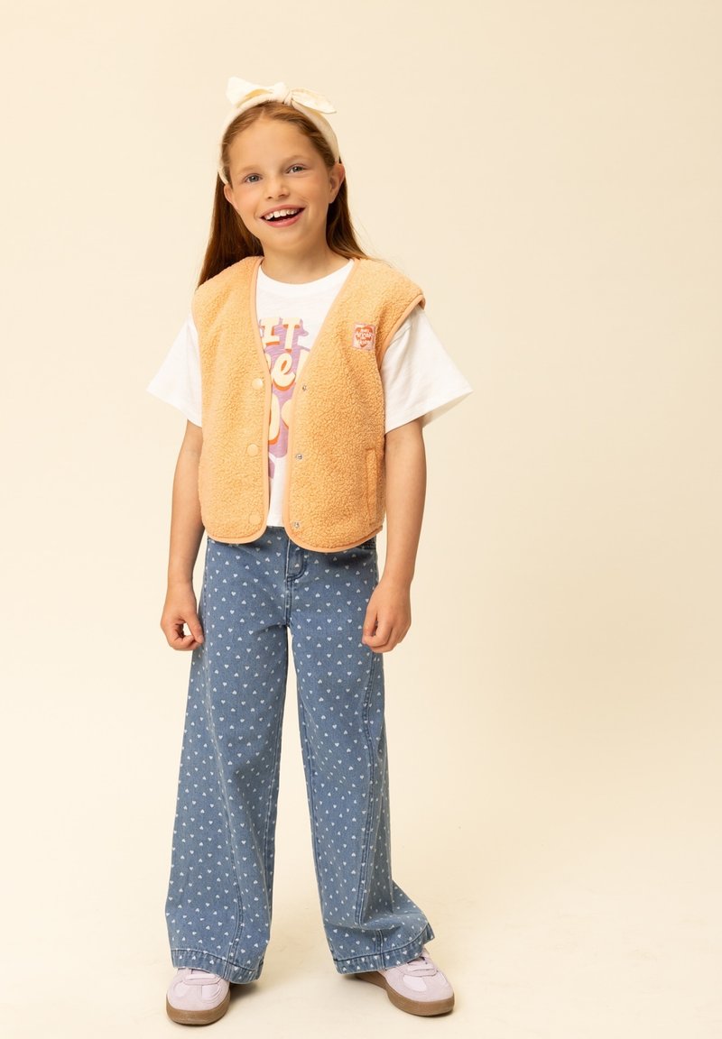 Oranje fleecevest met drukknopen over een wit T-shirt met print, gecombineerd met blauwe jeans met wijde pijpen en witte hartjespatronen, en lichte sneakers.