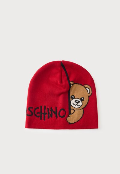Bonnet en tricot rouge avec un ours en peluche qui regarde derrière une ligne noire et un texte noir partiellement visible.