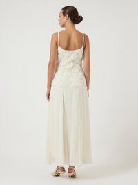 Γυναίκα που στέκεται με την πλάτη στραμμένη, φορώντας ένα κρεμώδες απογευματινό φόρεμα χωρίς μανίκια με λουλουδάτη δαντέλα στο μπούστο και ρέουσα φούστα, σε συνδυασμό με κρεμώδεις ψηλές γόβες.