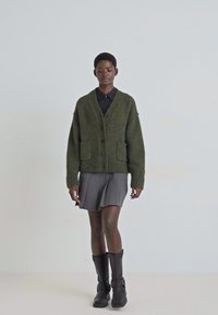 Groene gebreide cardigan met knopen, twee voorzakken, geribde manchetten, gedragen over een zwart shirt en grijze rok, gecombineerd met zwarte laarzen.