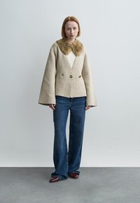 Μπεζ μάλλινο παλτό με τεχνητό γούνινο γιακά, διπλό κούμπωμα, μανίκια σε σχήμα καμπάνας, συνδυασμένο με σκούρο μπλε τζιν καμπάνα και καφέ παπούτσια.