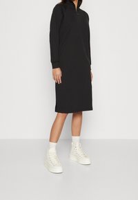 Schwarzes Langarmkleid mit Halbreißverschluss-Kragen, seitlichen Schlitzen, aus weichem Stoff. Kombiniert mit weißen High-Top-Sneakern und gerippten Socken.