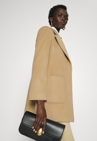 Beige wollen jas met brede revers en een voorzak, gedragen met een zwarte leren clutch met een gouden ovale sluiting. Soepele textuur, op maat gemaakt.
