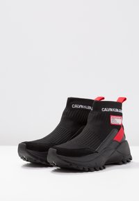 Černé tenisky v ponožkovém stylu s robustními podrážkami, červené poutka na patě, logo Calvin Klein Jeans na manžetě a bočním nápletu.