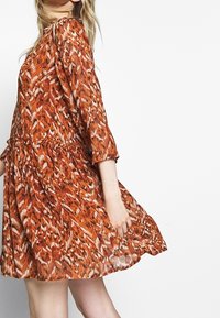 Femme portant une robe orange ample jusqu'aux genoux avec un motif, manches trois-quarts et taille froncée.