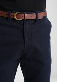 Blå bomullsbukser med en slät textur, utrustade med ett brunt läderbälte och en mässingsspänne. Fickor med subtila sömnadsdetaljer.