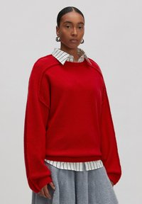 Roter Pullover mit lockerer Passform, gerippten Bündchen und rundem Halsausschnitt, über einem weißen gestreiften Hemd mit Kragen getragen, kombiniert mit einem grauen Rock.
