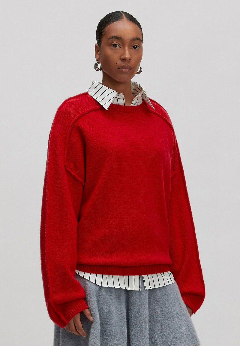 Roter Pullover mit lockerer Passform, gerippten Bündchen und rundem Halsausschnitt, über einem weißen gestreiften Hemd mit Kragen getragen, kombiniert mit einem grauen Rock.