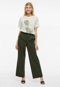 Valkoinen lyhythihainen paita, jossa on vihreä lehtikuosi, yhdistettynä korkeavyötäröisiin, leveälahkeisiin oliivinvihreisiin housuihin. Mustat avonaiset kengät.