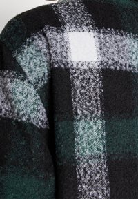 Uldstof med stort ternet mønster i grønne, sorte, hvide og grå toner, der viser teksturerede vævede detaljer.