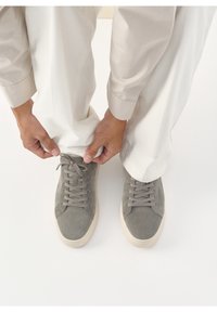 Personne en pantalon crème ajustant le poignet au-dessus de baskets en daim gris avec semelles beiges sur un sol blanc.