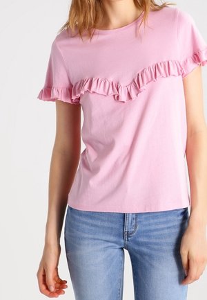 Vrouw draagt een lichtroze T-shirt met korte mouwen en diagonale ruches en blauwe spijkerbroek, staande voor een effen achtergrond.