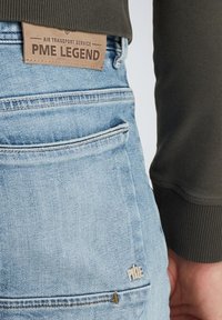 Hellblaue Denim-Jeans mit einem braunen Lederetikett, auf dem "PME LEGEND" steht. Die Merkmale umfassen eine Gesäßtasche mit besticktem Logo und sichtbaren Nähten.