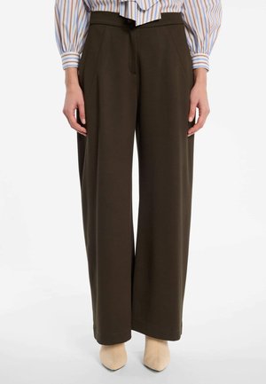 Vrouw draagt donkerbruine broek met wijde pijpen, beige puntige schoenen en een gestreepte blouse met lange mouwen en pofmouwen.