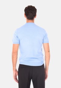 Uomo in piedi di spalle, vestito con una camicia a maniche corte azzurro chiaro e pantaloni neri, davanti a uno sfondo bianco semplice.