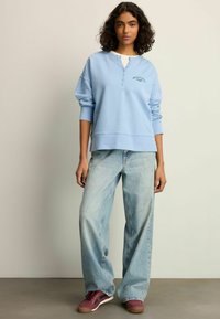 Blå langærmet pullover med knaplukning, broderede detaljer, parret med let vaskede vide jeans og bordeaux sneakers.