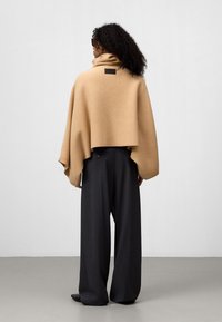 Poncho color cammello con colletto alto, realizzato in tessuto morbido, con una patch del logo sul retro, abbinato a pantaloni grigi scuri dal taglio ampio.
